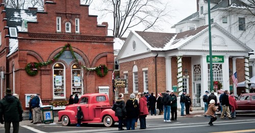 A vintage red truck parked near historic buildings, with people gathered outside in a snowy town setting.