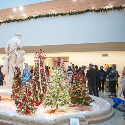 A festive scene with decorated Christmas trees around a statue, and a crowd enjoying the holiday display in a museum.