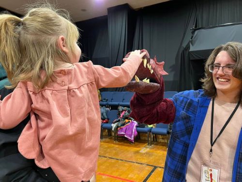A child interacts with a puppet while a smiling adult watches, both in a theater setting.