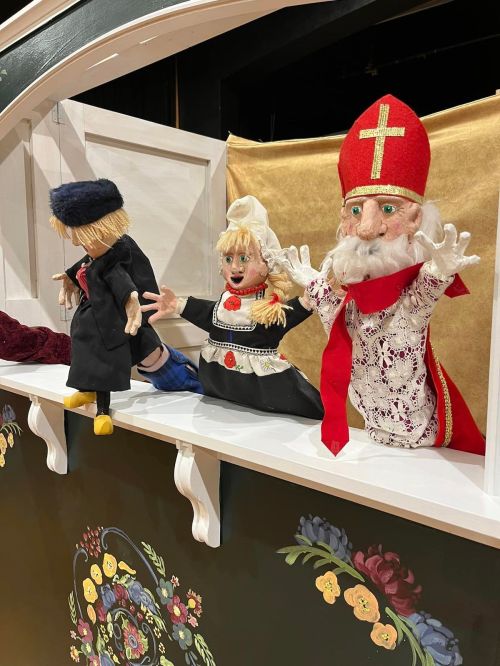 Three colorful puppets on a stage: a boy in a hat, a girl in a dress, and a bearded figure in a red hat.