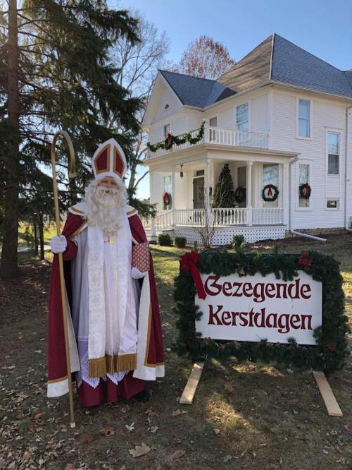 A man dressed as Santa stands beside a festive sign reading "Gezegende Kerstdagen" in front of a decorated white house.