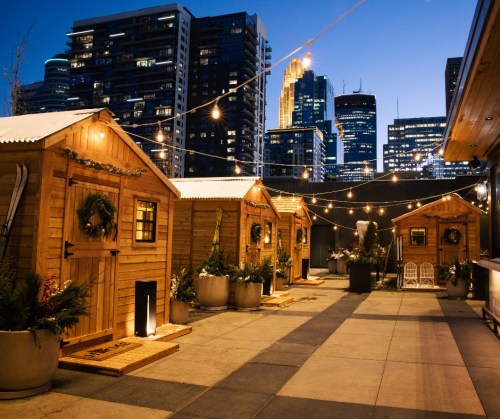 Cozy wooden cabins with festive decorations, illuminated by string lights, against a city skyline at dusk.