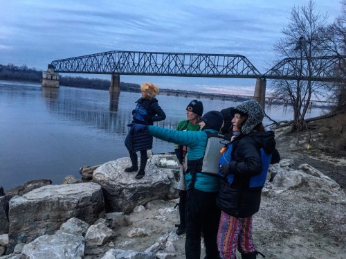A group of people in life jackets stand on rocks by a river, pointing towards a bridge in the distance at dusk.