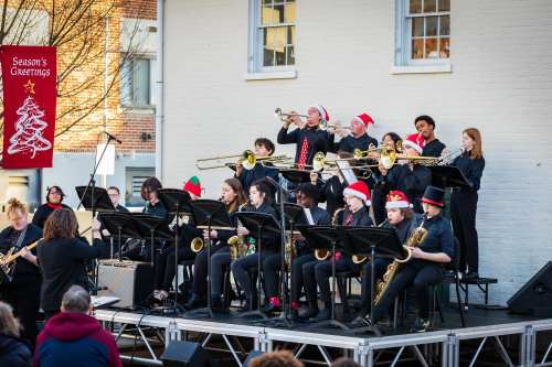 A festive band performs outdoors, wearing holiday hats, with a conductor leading the musicians on a stage.