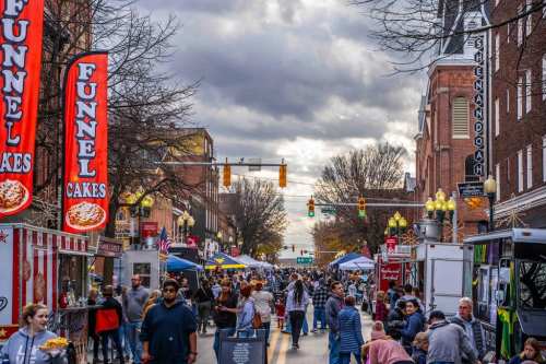 A bustling street fair with food vendors, colorful banners, and a crowd enjoying the festivities under a cloudy sky.
