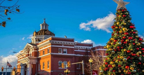 A festive Christmas tree adorned with ornaments stands in front of a historic building under a bright blue sky.