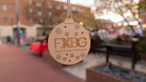 A wooden ornament with "FXBG City of Fredericksburg, VA" and snowflake designs, hanging outdoors.