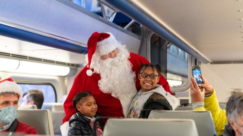Santa Claus poses with two children on a train, while other passengers smile and take photos.