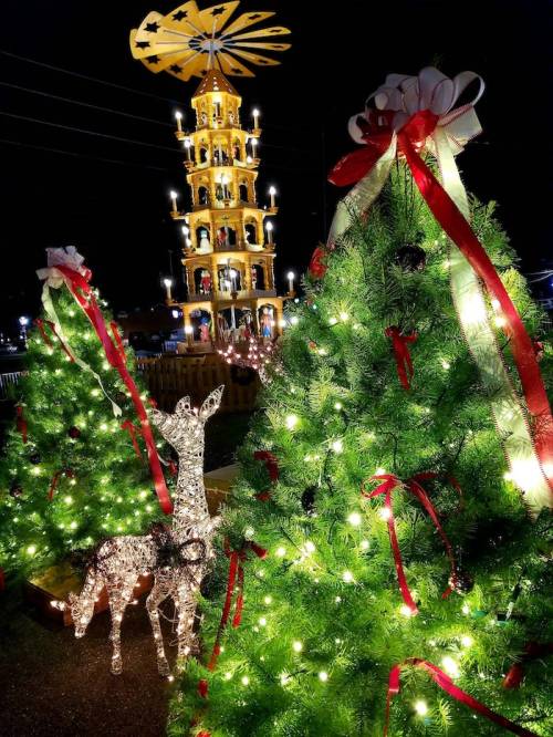 A festive scene featuring illuminated Christmas trees, a decorative deer, and a tall, brightly lit tower at night.