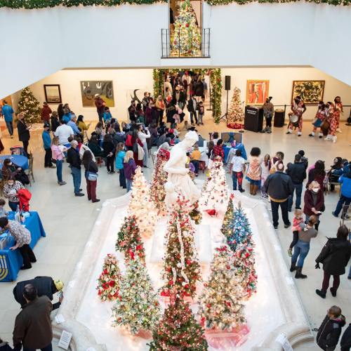 A festive indoor scene with a large crowd, decorated Christmas trees, and a central fountain, all in a gallery setting.