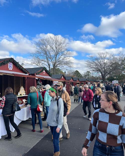 A bustling outdoor market with people browsing stalls under a blue sky with clouds. Trees are visible in the background.
