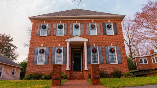 A brick house with festive wreaths on the windows, surrounded by greenery and trees, under a cloudy sky.