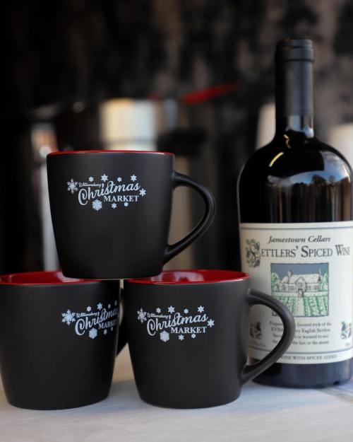 Three black mugs with red interiors featuring "Christmas Market" text, alongside a bottle of spiced wine.