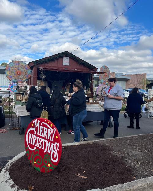 A festive market scene with people shopping at a booth decorated for Christmas, featuring a "Merry Christmas" sign.