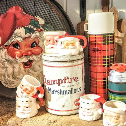 A festive display featuring Santa-themed mugs, a vintage marshmallow tin, and a plaid thermos on a wooden surface.