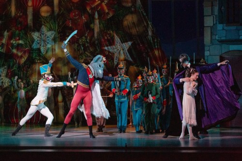 A theatrical performance featuring dancers in colorful costumes, including a soldier, a character with a horse head, and a girl.