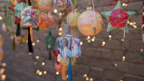 Colorful decorative ornaments hang against a brick wall, illuminated by soft fairy lights.