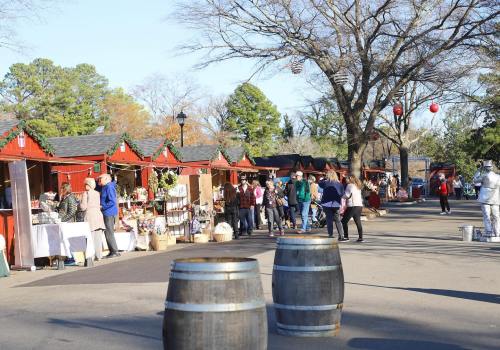 A bustling outdoor market with wooden stalls, shoppers, and festive decorations under clear blue skies.