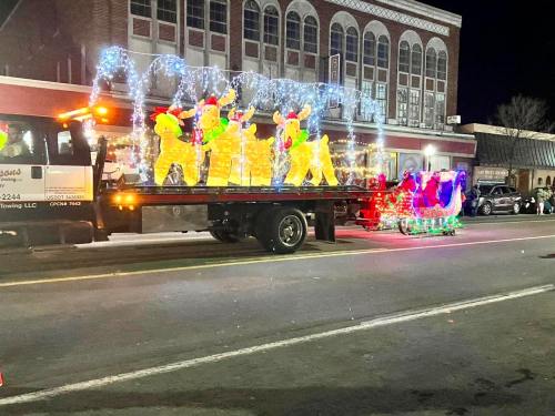 A festive parade float featuring illuminated reindeer and a colorful sleigh, decorated with lights, on a city street at night.