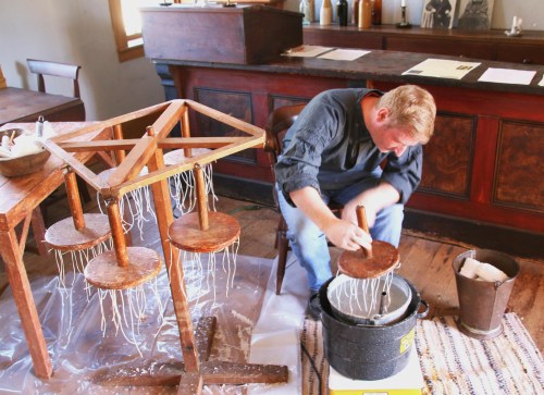 A person works on a candle-making process, using a wooden frame and dipping wicks into a container of wax.