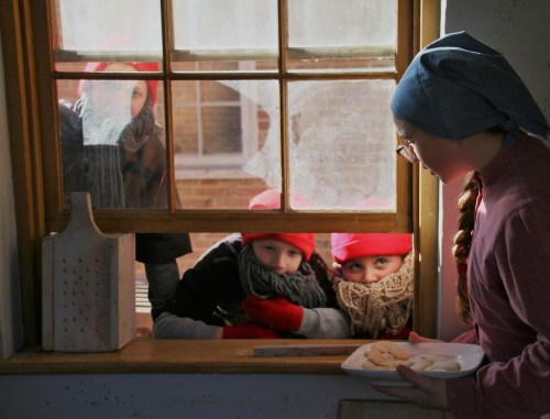 Children in red hats peek through a window while a girl in a headscarf serves cookies inside.