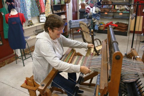 A woman weaves on a loom in a craft shop, surrounded by colorful fabrics and handmade items.