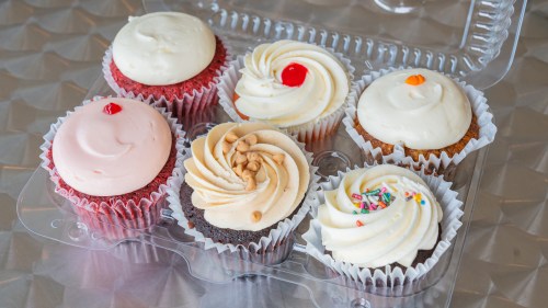 A clear container holds six colorful cupcakes with various frosting designs and toppings.