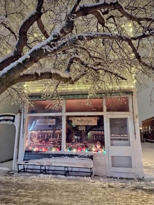 A cozy, snow-covered storefront with colorful lights and a tree overhead, creating a warm winter atmosphere.