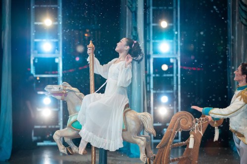 A performer in a white dress joyfully rides a carousel horse under bright stage lights, with snowflakes falling around.