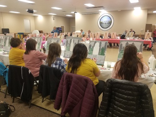 A group of people painting on canvases in a community hall, with easels set up in front of them.