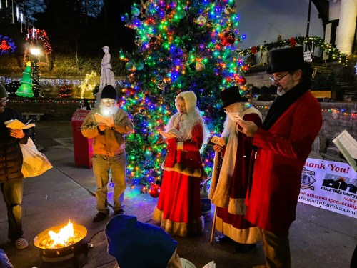 A group of people in festive attire sings carols by a brightly lit Christmas tree and a small fire.