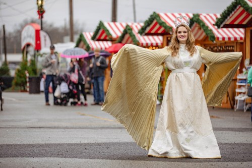 A woman in a golden gown and crown poses in a festive market with holiday decorations and people in the background.