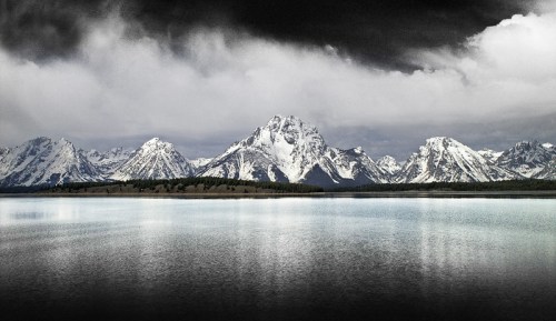 A serene lake reflects snow-capped mountains under a cloudy sky, creating a dramatic and tranquil landscape.