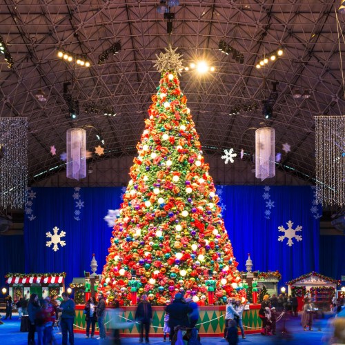 A large, colorful Christmas tree adorned with ornaments, surrounded by festive decorations and people in a holiday setting.