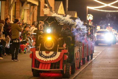 A festive train with smoke puffs travels down a street, surrounded by cheering spectators and holiday lights.
