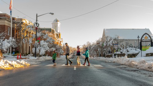 A family of four crosses a snowy street in a quaint town, with snow-covered buildings and trees in the background.