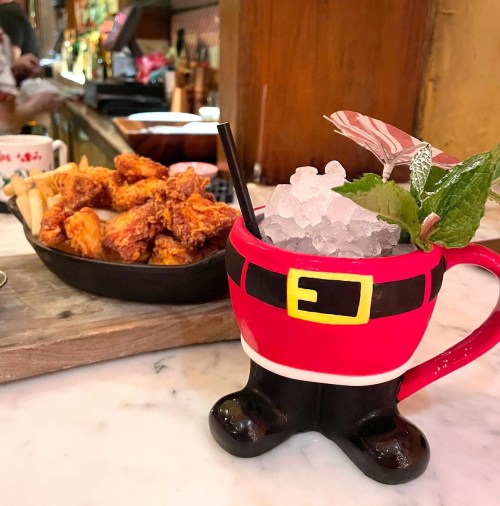 A festive drink in a Santa-themed mug with ice, alongside a plate of crispy chicken wings on a marble table.