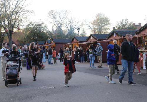 A bustling outdoor market scene with people browsing stalls, children playing, and festive decorations in the background.
