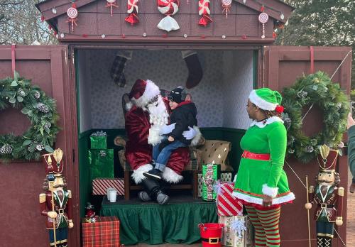 A child sits on Santa's lap in a festive setting, while an elf stands nearby, surrounded by holiday decorations and gifts.