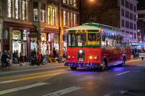 A festive, brightly lit trolley bus decorated with holiday lights drives past a crowd on a city street at night.