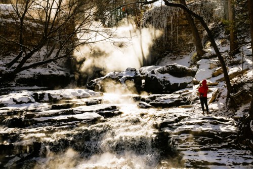 A person in a red hoodie stands by a snowy waterfall, surrounded by mist and icy trees in a serene winter landscape.