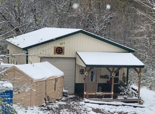 A snow-covered cabin with a green roof, porch, and a nearby tent in a winter forest setting.