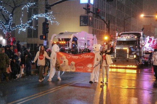 Two people in polar bear costumes hold a "Happy Holidays" banner during a festive parade with a truck in the background.