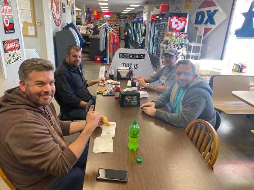 Four men sitting at a table in a casual setting, enjoying food and drinks, with various items in the background.