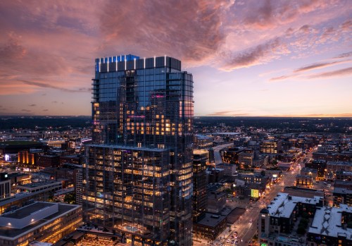 A modern skyscraper illuminated at dusk, surrounded by a city skyline under a colorful sunset.