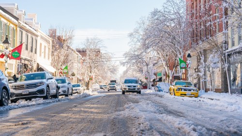 A snowy street lined with cars and decorated with festive flags, under a clear blue sky.