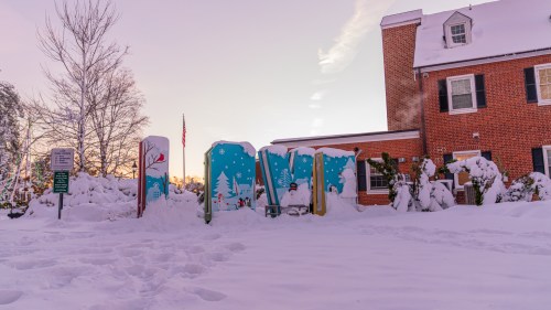 A snowy scene featuring colorful play structures partially buried in snow, with a brick building and trees in the background.