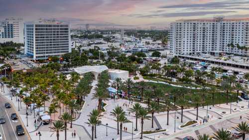 Aerial view of a waterfront park with palm trees, people, and boats, surrounded by buildings and a colorful sunset sky.