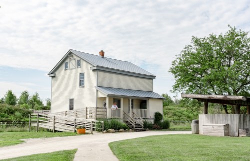 A two-story yellow house with a metal roof, surrounded by greenery and a gravel path, under a cloudy sky.