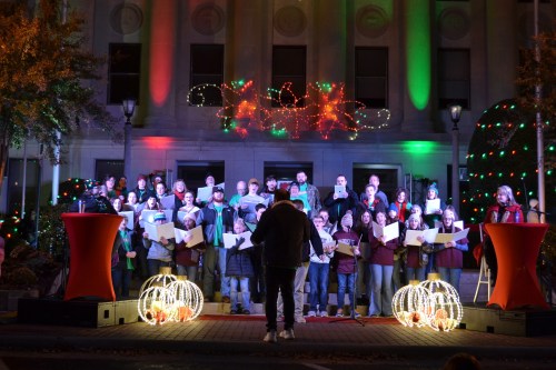 A choir performs outdoors in front of a decorated building with festive lights, surrounded by holiday decorations.